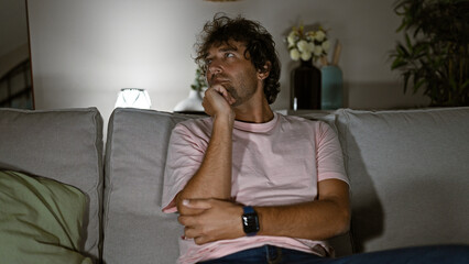 Fototapeta premium Pensive young man sitting on a couch in a dimly lit living room, evoking a sense of contemplation and comfort.