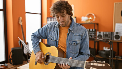 Handsome hispanic man playing guitar in a music studio, showcasing creative vibe and artistic focus.