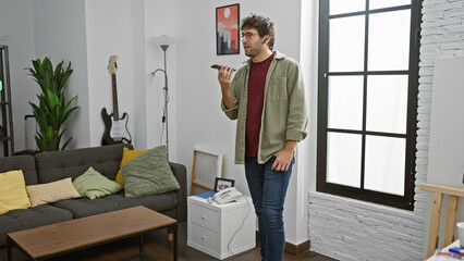 A casual young man with a beard sending a voice message indoors, standing in a modern living room...
