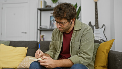 Focused hispanic man writing in notebook on sofa in a cozy living room with guitar