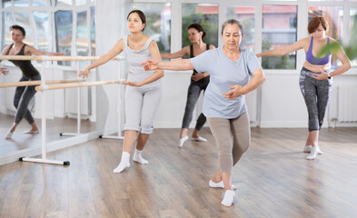 Poised senior woman, dressed in comfortable t-shirt and leggings, executing dance moves with focused precision among group of younger female ballet amateurs rehearsing in studio..