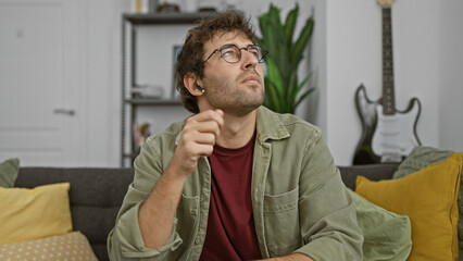 Young hispanic man with beard and glasses wearing wireless earbuds at home, contemplating while...
