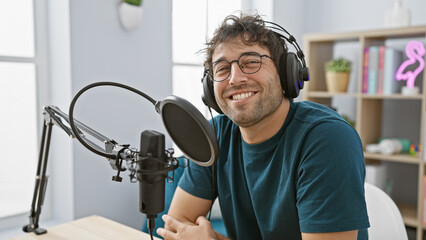 Smiling young hispanic man with headphones operating a microphone in a bright indoor studio.
