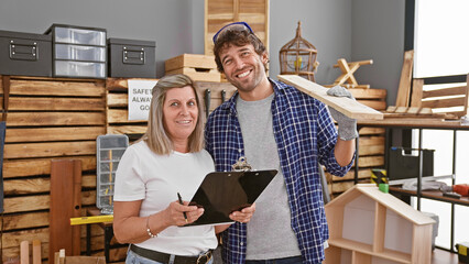 Two smiling carpenters, man and woman, confidently taking notes on clipboard while holding wood...