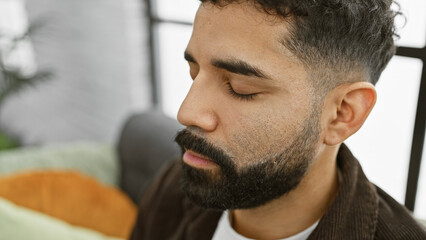 A handsome young adult hispanic man with a beard poses indoors, eyes closed in a serene, home environment.