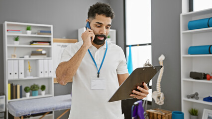 A young hispanic man with a beard is working indoors at a clinic, talking on the phone and holding...
