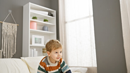 Blonde toddler boy sitting thoughtfully on a bed inside a well-lit, cozy bedroom.