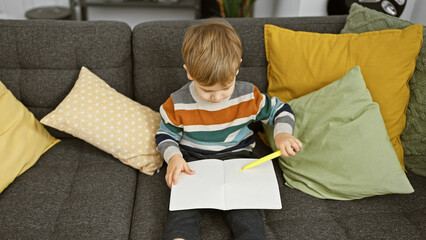 Toddler boy drawing in a notebook on a couch indoors surrounded by colorful pillows.