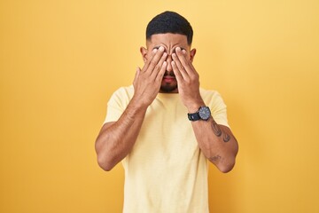 Young hispanic man standing over yellow background rubbing eyes for fatigue and headache, sleepy...