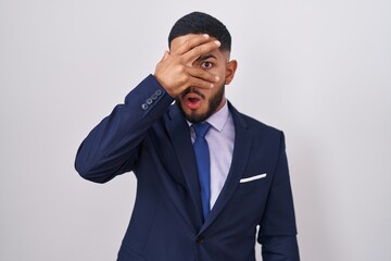Young hispanic man wearing business suit and tie peeking in shock covering face and eyes with hand, looking through fingers with embarrassed expression.