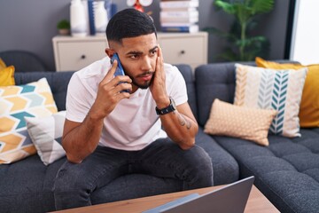 Young latin man talking on smartphone with worried expression at home