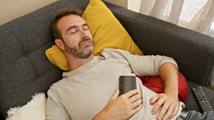 A middle-aged man with beard sleeps on a sofa holding a phone, encapsulating rest in a cozy living...