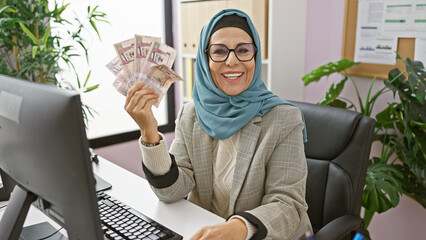 A mature woman in a hijab smiling while holding saudi riyals in a modern office interior.