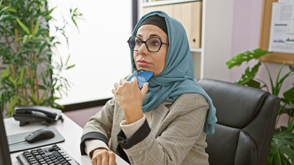 A middle-aged woman in hijab listens intently on a smartphone in her modern office space, surrounded by indoor plants and technology.
