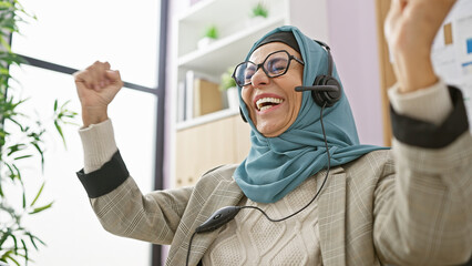 A jubilant middle-aged woman wearing a hijab celebrates in an office setting with headphones on.