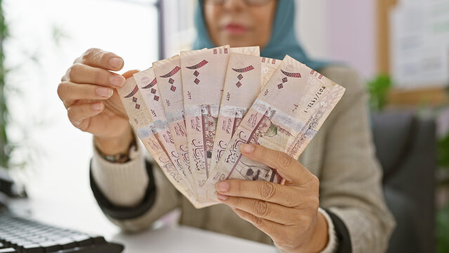 Middle-aged woman counting saudi riyal banknotes at her office desk, indicating finance and business in a workplace setting.