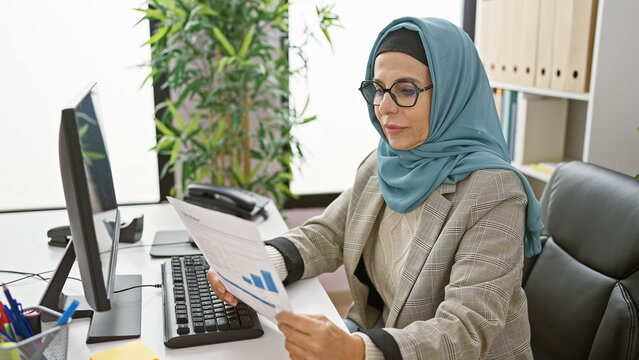 A mature muslim woman reviews financial documents in a modern office setting, exuding professionalism and focus.