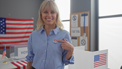 A smiling blonde woman in a striped shirt points to her 'i voted' sticker in a us polling station...