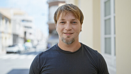 Portrait of a smiling young blond man with a beard, wearing a casual dark shirt, standing on a city...