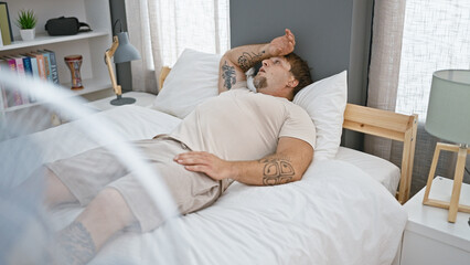 A young bearded man lounging in bed, with tattoos visible, inside a modern bedroom subtly hinting...