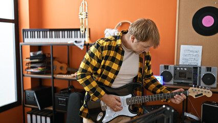 A blond man plays electric guitar in a music studio filled with various instruments and recording...