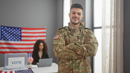 Smiling soldier in uniform crossing arms in front of a us flag and voting booth with a female...