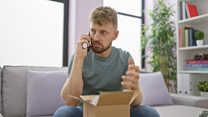 Caucasian man with blue eyes and beard talking on phone while unpacking a box in his cozy living...