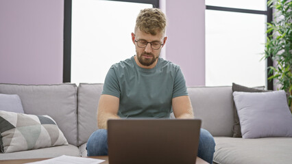 A focused young man with a beard and glasses using a laptop on a couch in a modern living room.