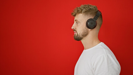 Profile of a young caucasian man with blue eyes and a beard, wearing headphones against a red...