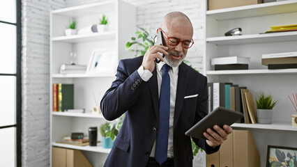 A bearded, mature man in a suit converses on the phone while holding a tablet in a modern office...