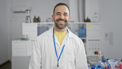 Portrait of a smiling man with a beard, wearing a lab coat and id badge in a laboratory setting.