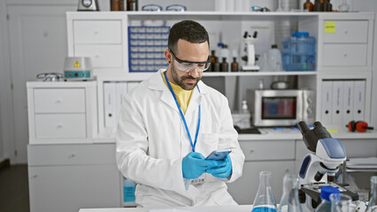 Hispanic man with beard in white lab coat examines a smartphone in a clinical laboratory setting, representing modern healthcare technology.