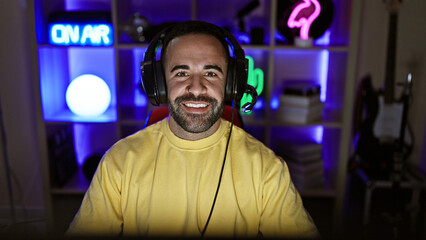 A smiling bearded hispanic man wearing headphones in a colorful gaming room at night.