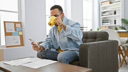 Hispanic man with beard drinking coffee and checking smartphone in bright modern office.