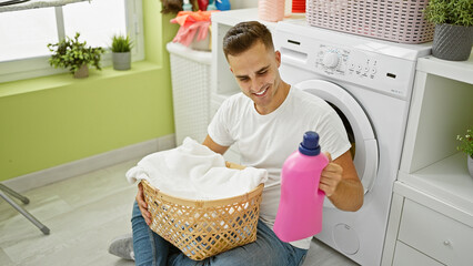 Handsome young man smiling while holding a laundry basket and detergent in a bright home laundry...