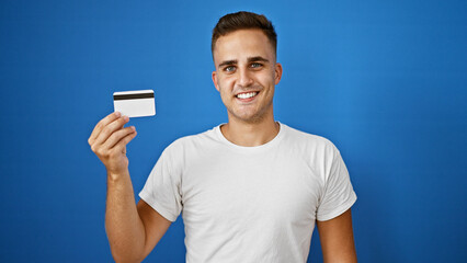 Handsome young hispanic man holding a credit card smiling against a blue background.