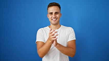 A cheerful young man clapping hands against a vibrant blue isolated background, embodying joy and...