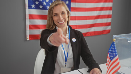 Smiling woman points at camera in a voting center with american flag, showing civic participation.