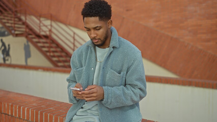 Handsome black man in casual attire using a smartphone on city stairs outdoors.