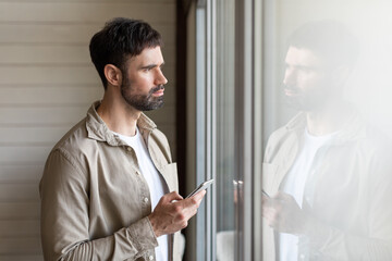 A man is standing in front of a window, looking at his cell phone. He appears focused on the...