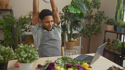 Handsome man stretching inside a floral shop with laptop and fresh flowers on desk, portraying a...