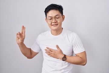 Young asian man standing over white background smiling swearing with hand on chest and fingers up,...