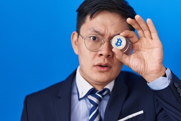 Young asian man holding virtual currency bitcoin covering eye in shock face, looking skeptical and sarcastic, surprised with open mouth