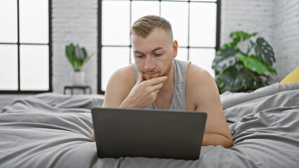 A thoughtful young man with a beard works on his laptop in a modern bedroom with large windows