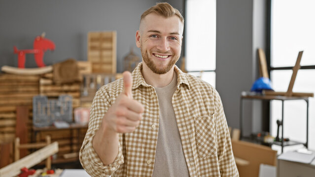 Handsome man with beard giving thumbs up in a carpentry workshop, surrounded by wood and tools.