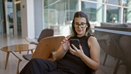 Radiant hispanic woman, all smiles, busily messaging on her smartphone while fashionably lounging...