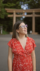 Fototapeta premium Cheerful beautiful hispanic woman with glasses, happily standing, looking around, enjoying the positive vibes of tokyo's meiji temple