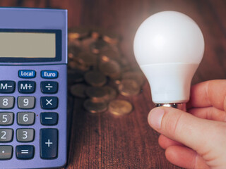 Glowing LED bulb in hand, calculator and stack of coins on wooden table. Cost of electricity for a...