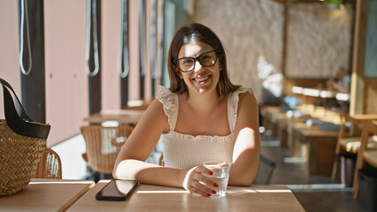 Beautiful young hispanic woman wearing glasses drinking a glass of water in a cafeteria