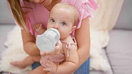 Loving mother giving a milk feeding bottle to her baby daughter while sitting in the relaxed comfort of their family home, cherishing an intimate indoor moment of infant nutrition and togetherness